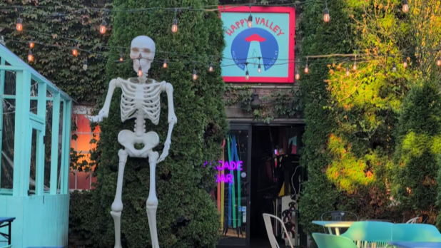 A large skeleton decoration stands in front of a greenery-covered building. Above the door, a sign reads "Happy Valley."