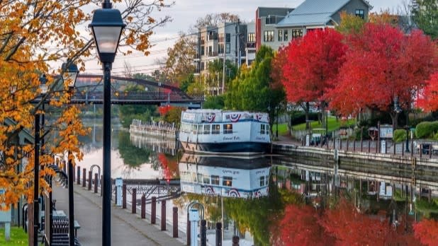 Autumn waterfront scene with the Colonial Belle on calm reflective water, colorful trees, lampposts, walkway, bridge, and buildings.