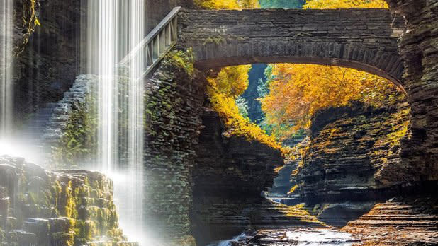 A waterfall to the left and bridge to the right in front of yellow fall leaves
