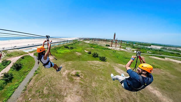 Two people zipline side by side along the sunny coast of Jones Beach at WildPlay Jones Beach; Photo Credit: @rmdragon on Instagram