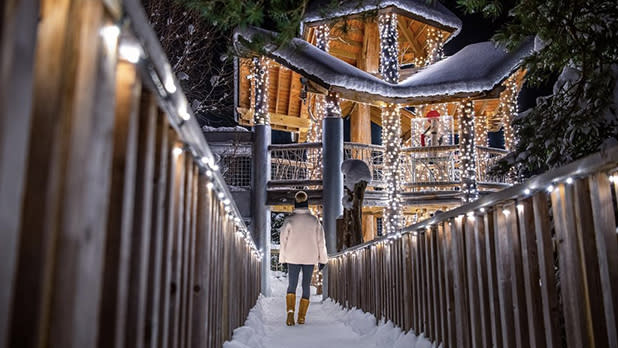 A person in a hat, white jacket, jeans, and tan boots walks down a snow-covered path toward a wooden treehouse covered in white lights