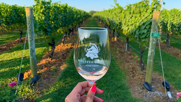 A woman with pink nails holding up an empty branded wineglass from Wolffer Estate Vineyard with views of the green vineyard in the sunlight