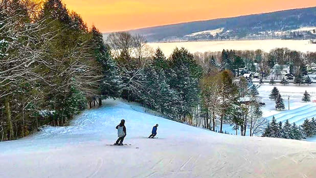 Two snowboarders traveling downhill past pine trees along the snowy slopes of Woods Valley Ski Area at sunset