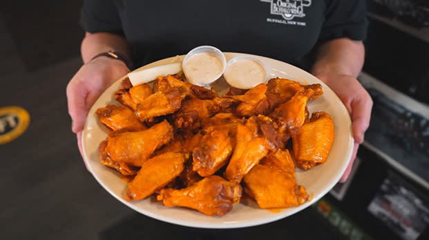 A person in a black t-shirt holds a plate of Buffalo wings with two dipping sauces