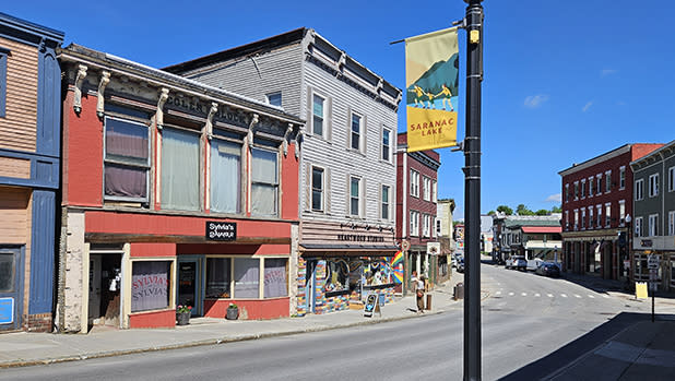 A quaint street in Saranac Lake with colorful historic buildings, a shop displaying vibrant artwork, and a clear blue sky. A banner reads "Saranac Lake."