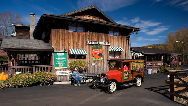 Photo of a man sitting on a bench next to a red vintage car in front of the Fly Creek Cider Mill