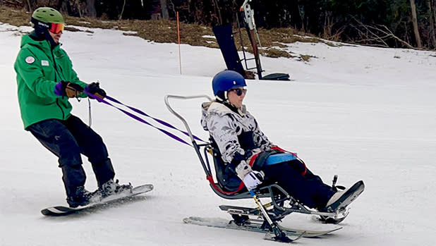 Nikki Walsh of Adapt Fit using adaptive skiing equipment on a snowy slope at Windham Mountain Club, assisted by an Adaptive Sports Foundation instructor in green.