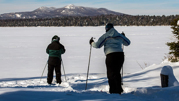 Two people dressed in winter gear Cross Country skiing on Lake Clear with mountains in the background.