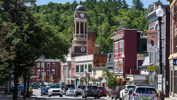 Saranac Lake's main street filled with parked cars, historic brick shops, clock tower, leafy hills, streetlamps, and colorful signs.
