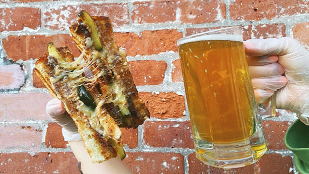 A person holds up a mug of beer and grilled cheese sandwich in front of a brick wall at Brew Cheese