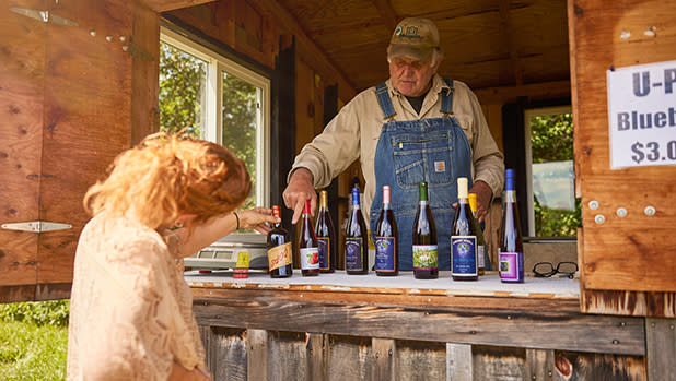 An man in overalls stands in a rustic wooden booth displaying colorful wine bottles. A woman selects a bottle, creating a warm, inviting scene at Roots & Rhythm.