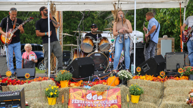 Live band performs on a colorful outdoor stage, with singer, guitars, drums, hay bales, flower displays, and a cheerful crowd at the Annual Festival of Colors in Wilmington.