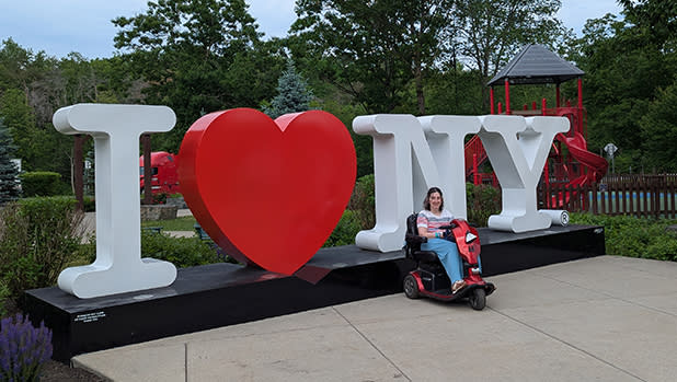 A person in a mobility scooter is smiling, posed beside a large "I LOVE NY" sign in front of the Adirondacks Welcome Center. Green trees and a playground are visible in the background.