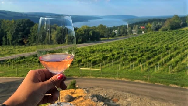 A person holds a glass of wine filled with rose with Keuka Lake in the background at the Living Roots Winery and Tasting Room