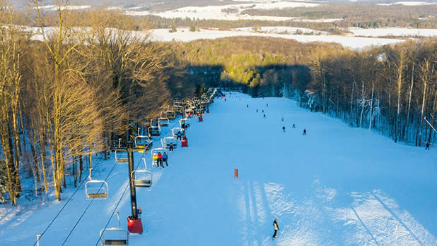 Aerial view of a snowy ski slope flanked by trees, with skiers descending. A line of ski lifts on the left and a bright, sunlit landscape in the distance.