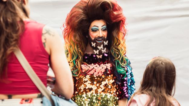 Performer in drag with rainbow hair, fabulous make-up, sequined rainbow outfit, and butterflies in their beard talking to a participant during Vancouver's annual Pride Block Party