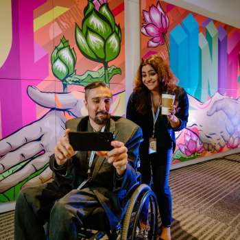A man and a wheelchair and a woman standing up pose for a selfie in front of a mural at the Greater Columbus Convention Center.