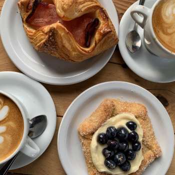 Top-down view of wooden table topped with plates of pastries and foam-art coffees at Fox In the Snow Cafe