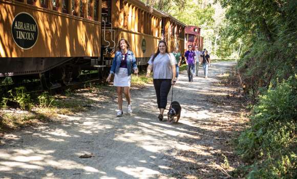 two people walking on rail trail with their dog next to northern central railway of york's steam engine