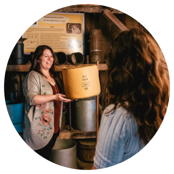 A museum tour guide holding a cheese wheel, showing a visitor around an 1800s replica factory
