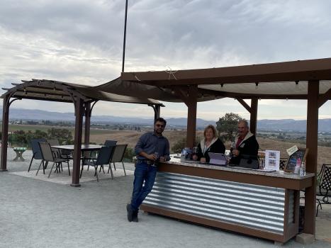 A guest stands at the open-air wine tasting counter at L&M Vineyard with the owners, with the view of Gilroy and the mountains in the a=background
