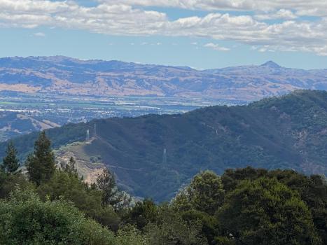Elevated view of Gilroy looking eastward from Mt. Madonna Summit, showing redwood covered mountain range, the city resting in the valley below and the Diablo mountain range in the distance with blu sky and clouds above.