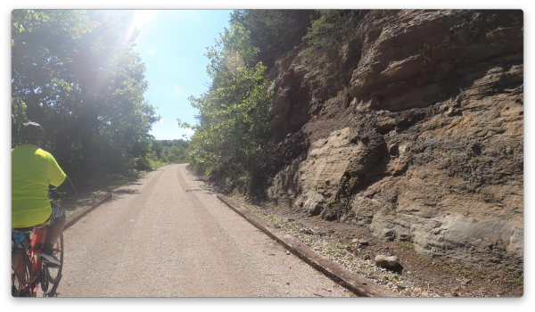 A cyclist in a bright yellow shirt rides on a gravel path beside a rocky cliff, surrounded by lush greenery and blue sky.