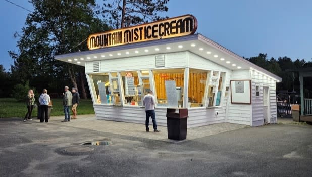 Ice cream stand at dusk, with bright neon "Mountain Mist Ice Cream" sign. Several people gather outside, creating a relaxed and nostalgic vibe.