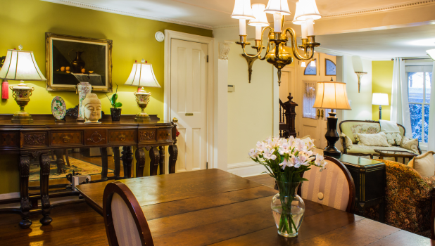 Elegant traditional dining room with a wood table, floral centerpiece, chandelier, antique console, lamps, and a sunlit sitting area inside the Arbor View House Bed and Breakfast.