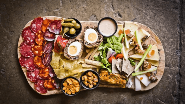 A huge charcuterie board with cured meats, crisps, cheese, a scotch egg, pickles, crackers and celery on a wooden serving board.