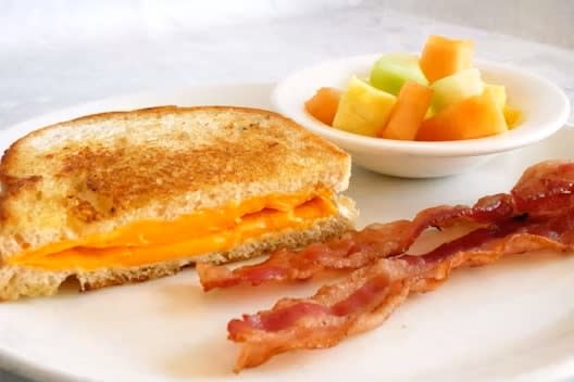 A grilled cheese sandwich with melted cheddar on a plate, accompanied by crispy bacon strips and a small bowl of cubed melon and cantaloupe.