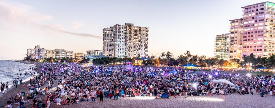 A large crowd sitting on a beach, facing north as they wait to watch a drone show when it gets dark