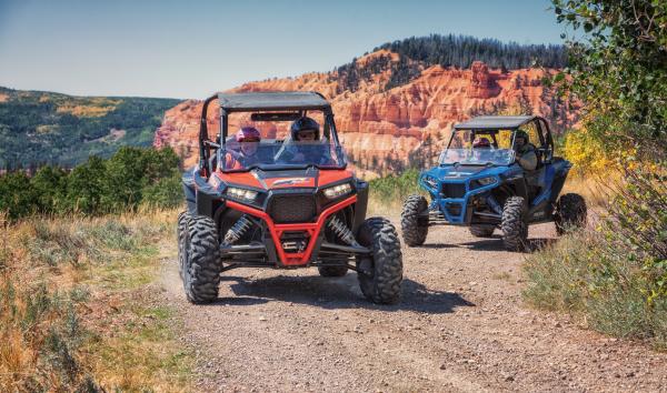 A red OHV followed by a blue OHV round the corner of a dirt road with pine trees and red rock formation in the background.