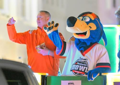 A mascot dressed as a blue dog in a football jersey and a football coach in an orange shirt wave to the crowd from a Mardi Gras float