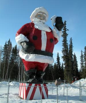 Large outdoor statue of Santa Claus in a traditional red suit with spruce trees and blue sky in background