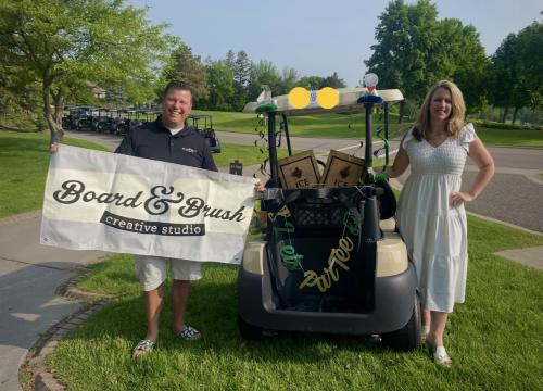 Board & Brush Woodbury Studio staff getting ready for Chamber golf tournament, holding a promotional sign next to a golf cart.