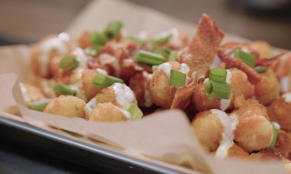 Image of a serving of potato tots served on a metal cookie sheet. The tots are topped with pieces of green scallions, bacon, and drizzled with a white sauce.