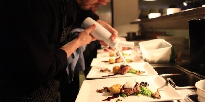 A chef in a dimly lit kitchen carefully plates gourmet dishes with precision, adding a final touch using a squeeze bottle. The atmosphere is focused and professional.