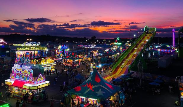Aerial view of carnival rides and games lit up as the sun sets casting shades of deep purple, orange and pink at the Great New York State Fair