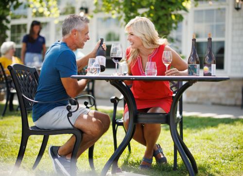 Couple laughing while sitting outside of Ledgestone Vineyards at a table and looking at a bottle of wine. There are two bottles of wine on the table and two glasses of wine.