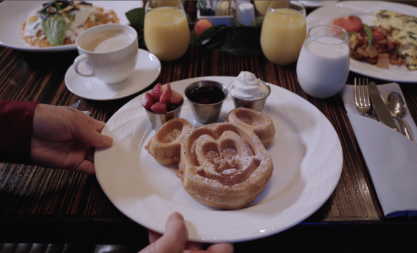 Image of a plate of waffles shaped like Mickey Mouse. Plate is placed on a dark brown/black wood table top. There is also a cup of coffee in the top left-hand corner, and a glass of milk and orange juice in the top right-hand corner. There are also two additional plates of food place in both top corners.