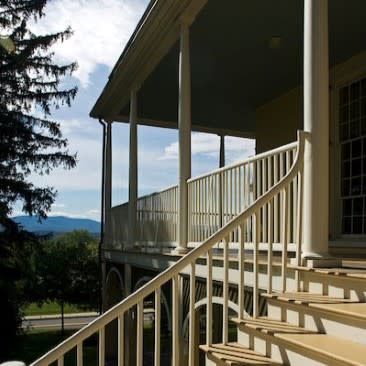 Covered porch with white railings and steps leads to Thomas Cole's historic home in Catskill, NY .  A large tree and mountains are visible in the background.