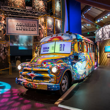 A brightly colored, psychedelic bus with vibrant patterns is displayed at an exhibition at Bethel Woods Center For The Arts. The sign reads "Aquarian Exposition," evoking a 1960s hippie vibe.