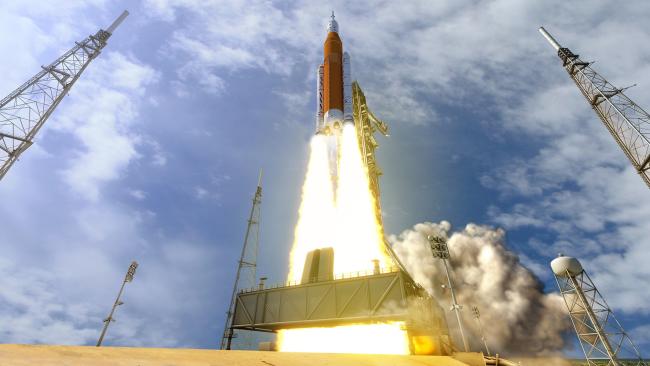 NASA's Space Launch System (SLS) rocket lifts off from the launch pad during the Artemis I mission, with bright flames and thick clouds of smoke marking the start of its journey into space beneath a partly cloudy sky.