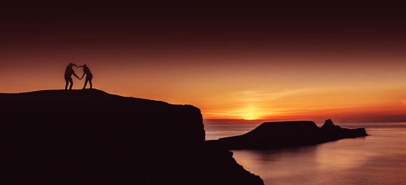 Two people making a heart with their arms with the backdrop of Worm's Head at sunset.