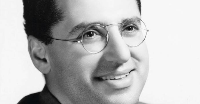 Black and white portrait of a smiling man wearing round glasses and a suit jacket.