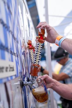 A hand pours beer from a decorative tap at a festive outdoor event, with a blurred crowd and colorful banners in the background.