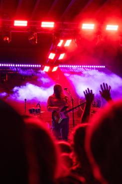 A musician plays bass on stage, surrounded by colorful lights and fog. Hands of the audience are raised, creating an energetic atmosphere.
