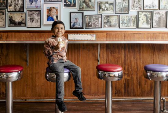 A young boy sits on an ice cream parlor stool with an ice cream cone in his hand.