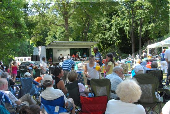 A crowd of people seated on lawn chairs listens to music on a stage.